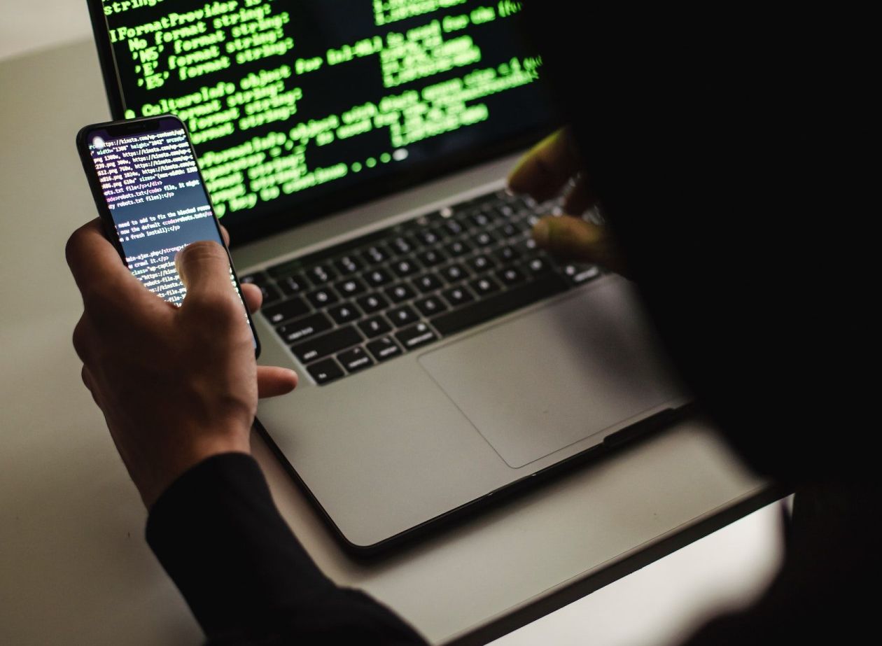 A photo of a man with a laptop on his lap holding a smartphone.